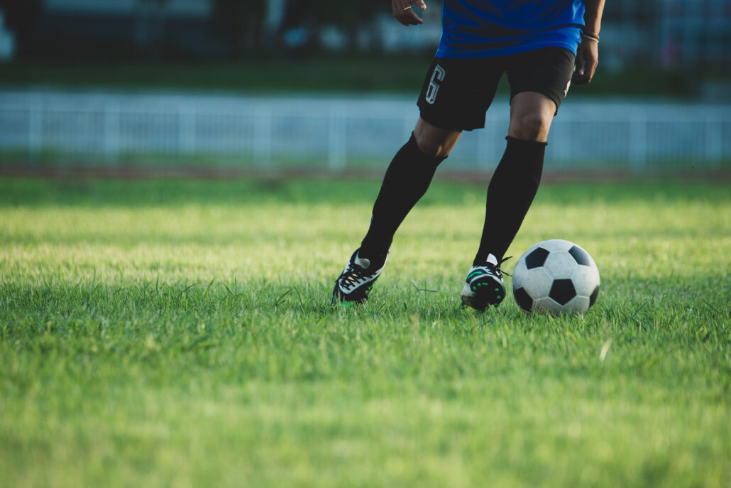 Plano médio inferior de jogador chutando bola em campo, representando as lesões esportivas de alta performance
